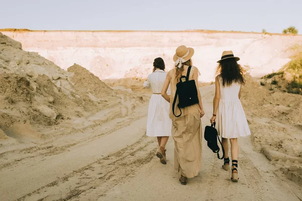 Back view of stylish girlfriends in straw hats walking in sandy canyon — Stock Photo