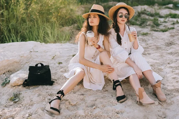 Привабливі дівчата в солом'яних капелюхах з кавовим латте, сидячи на землі разом — стокове фото