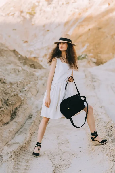 Красива дівчина в стильній сукні і солом'яному капелюсі позує з рюкзаком в піщаному каньйоні — стокове фото