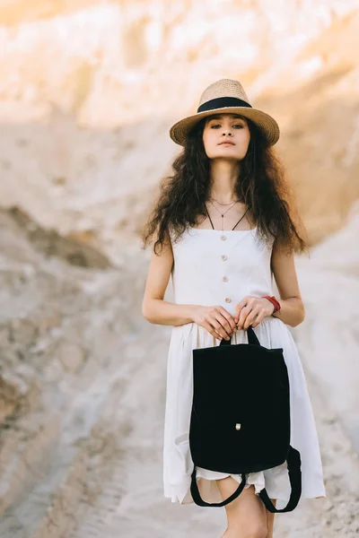 Стильная молодая женщина в соломенной шляпе держит сумку и позирует в песчаном каньоне — стоковое фото