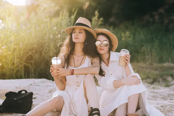 Belas amigas em chapéus de palha segurando copos de plástico com café latte e sentado no chão com luz solar — Fotografia de Stock