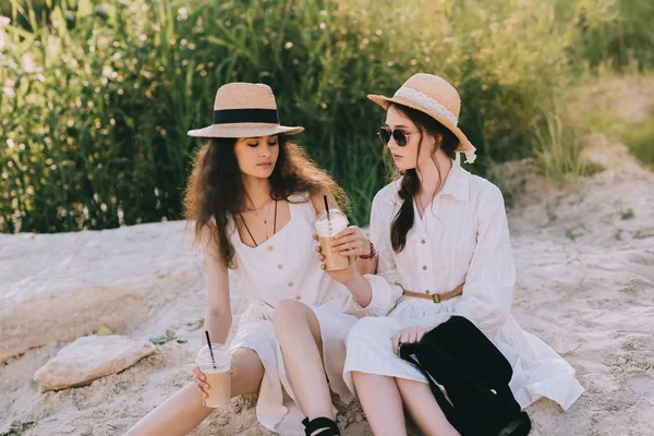 Привабливі подружки в солом'яних капелюхах з чашками кави лате сидить на землі — стокове фото