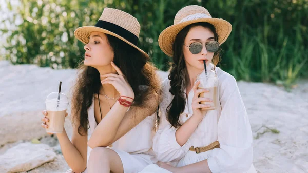 Beautiful female friends in straw hats drinking coffee latte and sitting on ground — Stock Photo