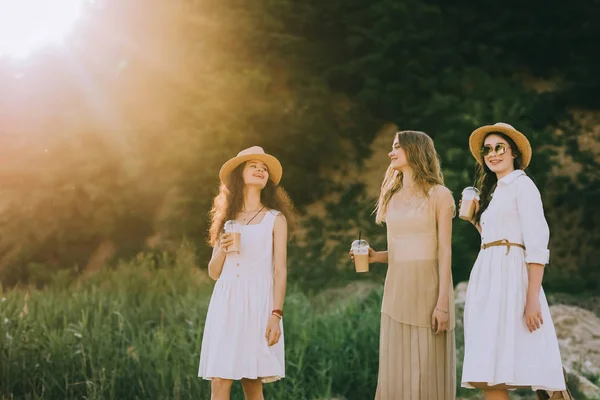 Belles amies en chapeaux de paille marchant dans la nature avec des tasses en plastique de café latte — Photo de stock