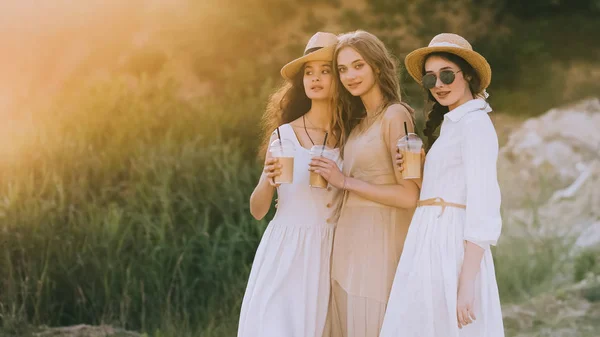 Chicas elegantes en sombreros de paja abrazando y sosteniendo café con leche, con luz solar - foto de stock