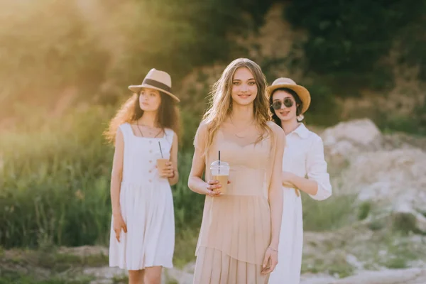 Chicas atractivas en sombreros de paja sosteniendo café con leche y caminando en la naturaleza con luz solar — Stock Photo