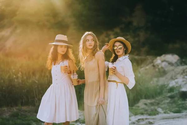 Elegante junge Frauen in Strohhüten, die Tassen mit Kaffeelatte in der Hand halten und mit Sonnenlicht posieren — Stockfoto