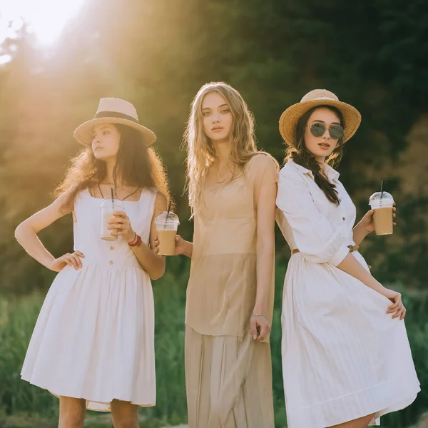 Meninas elegantes atraentes em chapéus de palha segurando café latte posando, com luz solar — Fotografia de Stock