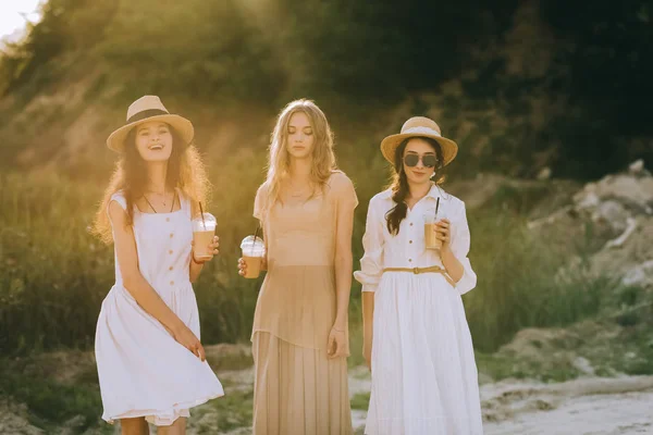 Meninas bonitas elegantes em chapéus de palha segurando xícaras de café latte e posando, com luz solar — Fotografia de Stock