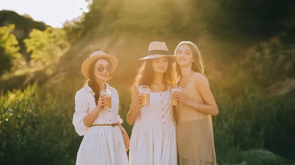 Élégantes copines élégantes dans des chapeaux de paille tenant des tasses avec café latte et posant, avec la lumière du soleil — Photo de stock