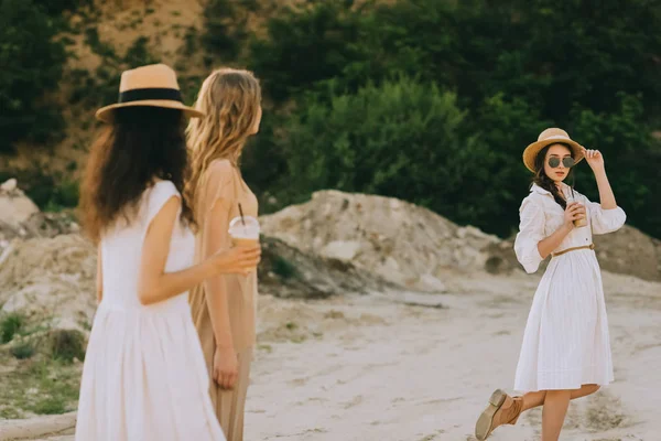 Chicas atractivas y elegantes en sombreros de paja caminando en la naturaleza con tazas de café con leche - foto de stock
