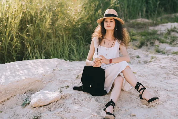 Atractiva chica rizada con café con leche y mochila sentado en el suelo - foto de stock