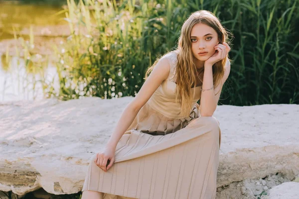 Attractive elegant girl in dress sitting on ground near pond — Stock Photo