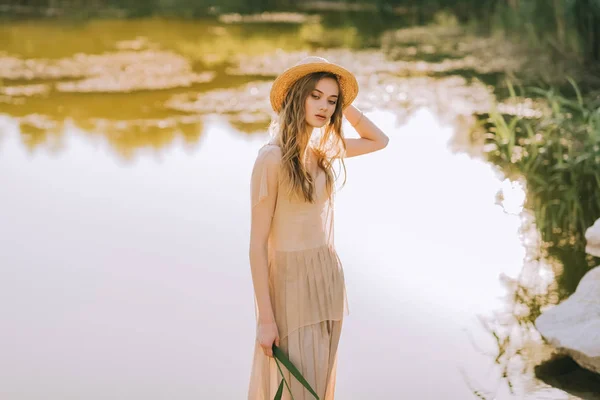 Hermosa chica rubia en vestido elegante y sombrero de paja posando cerca del lago - foto de stock