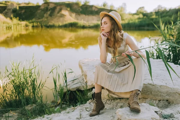 Chica con estilo en vestido elegante y sombrero de paja sentado en el suelo cerca del estanque - foto de stock