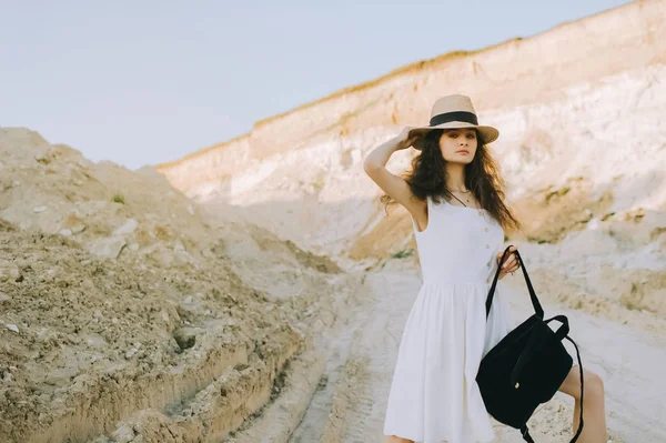 Beautiful curly girl in white dress and straw hat posing with backpack in sandy canyon — Stock Photo