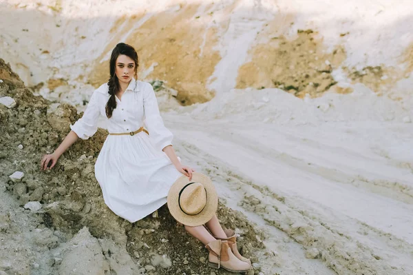 Beautiful girl in white dress with straw hat sitting in sandy canyon — Stock Photo
