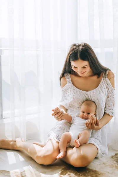 Щаслива молода мати сидить на підлозі біля вікна зі своєю чарівною маленькою дитиною вдома — стокове фото