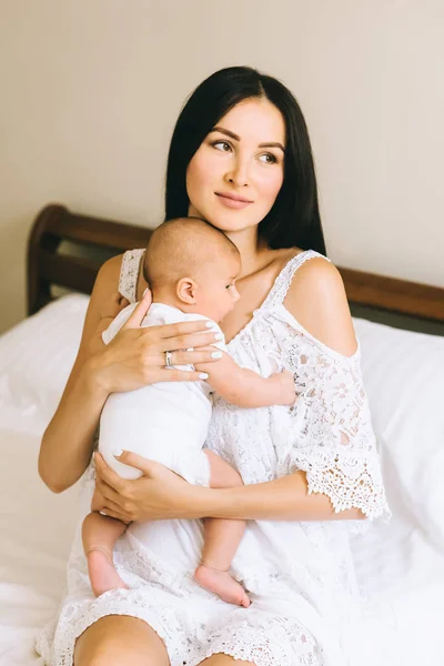 Feliz joven madre con su pequeño bebé sentado en la cama en casa - foto de stock