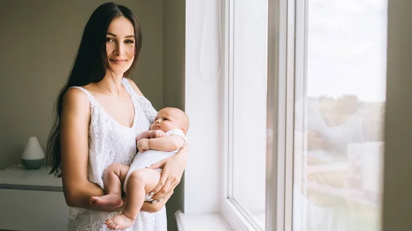Attraktive junge Mutter trägt ihr Baby am Fenster zu Hause und schaut in die Kamera — Stockfoto