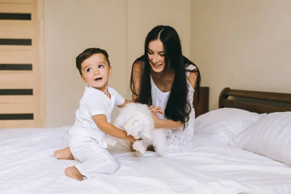 Glückliche junge Mutter und Kind spielen mit Hund Bichon, während sie zu Hause im Bett sitzen — Stockfoto