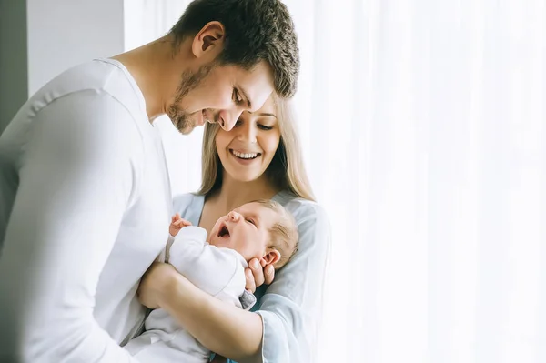 Sonriente familia llevando pequeño bebé niño delante de cortinas en casa - foto de stock