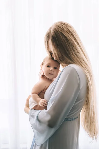 Hermosa madre llevando niño pequeño delante de las cortinas en casa - foto de stock