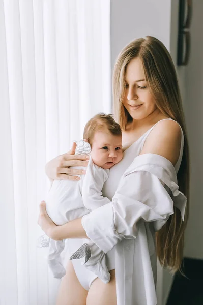 Sonriente madre llevando niño pequeño delante de las cortinas en casa - foto de stock