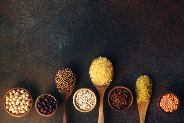 Различные органические злаки и злаки в деревянных ложках и чашах на темном каменном фоне
