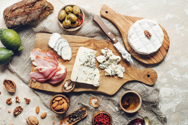 Set of cheese, hamon, honey, green olives, nuts and spices on rustic wooden board, variety of snacks on light concrete table, top view