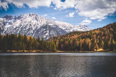 Karla kaplı dağda sonbahar renkli bir orman ve gölde bir yansıma var.
