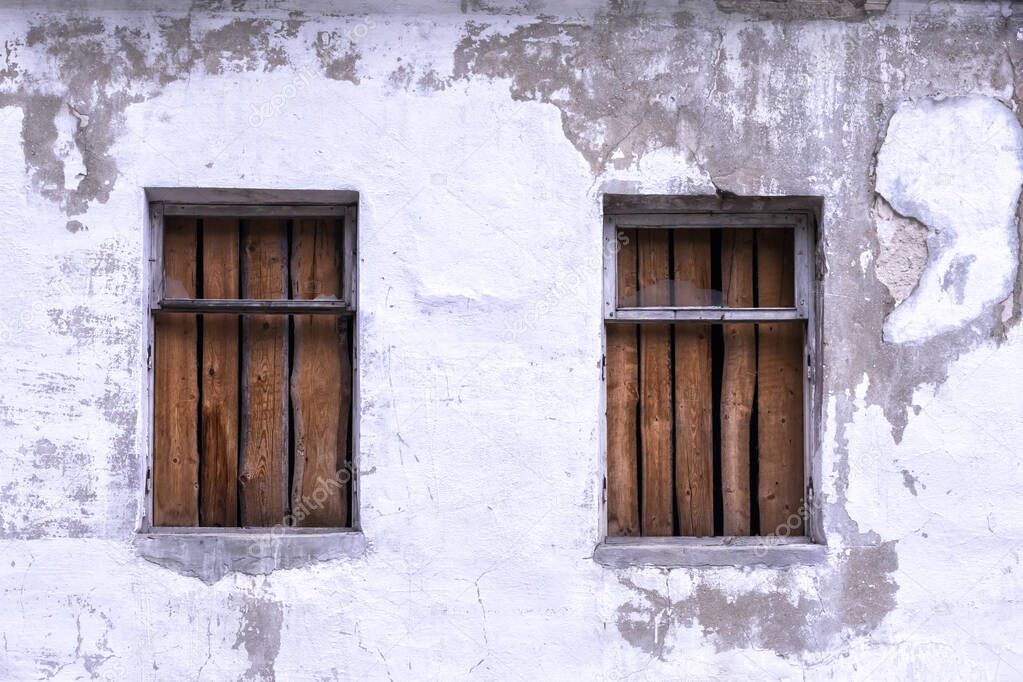 Two windows boarded up by wooden planks in a shabby plastered wall of an old building close up