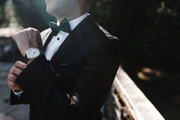 Man in black jacket, white shirt and green bow-tie clamps the watch on his arm.