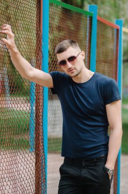 Tek eliyle çit yaslanmış güneş gözlüklü bir adam portresi.