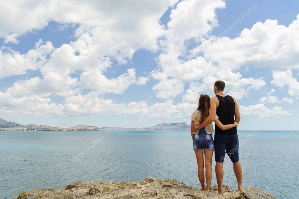A young man and woman standing at the rock and looking at the sea photographed from behind.