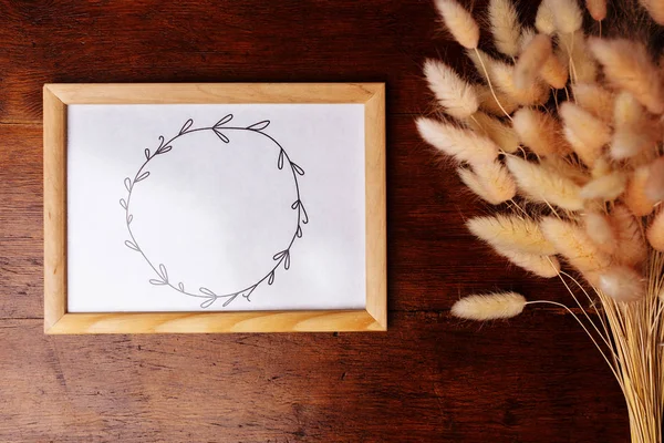 Painted wreath on the paper in the frame and flowers on the brown wooden background.