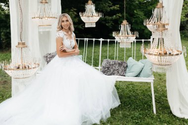 Gülümseyen gelin doğada bankta oturuyor ve ellerini göğsüne yaklaştırıyor. Gelin portresi