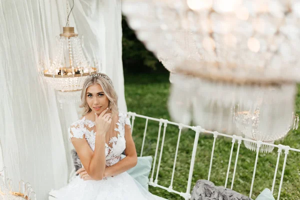 美しいブロンドの花嫁は白いベンチに座って手で彼女の顔に触れています 花嫁の肖像 — ストック写真
