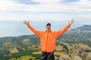 Turuncu ceketli bir adam, deniz manzaralı dağların tepesinde kolları göğe yükselmiş ve mutluluktan bağırıyor. Erkek portresi. Gerideki adam.