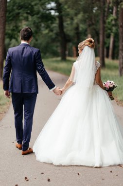 Gelinlik, duvak ve takım elbiseli damat el ele tutuşarak parkta yürüyorlar.
