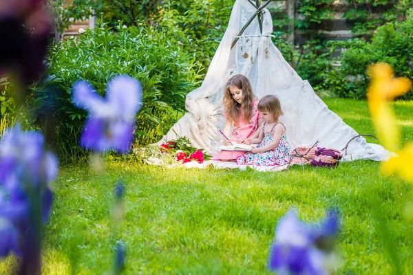 Two sisters in multi-colored dresses sit in white boho tent outd — Stock Photo, Image