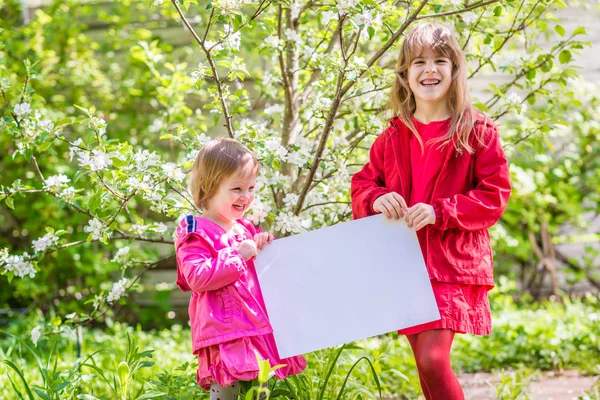 Two laughing sisters stand near the apple tree and hold a white — Stock Photo, Image