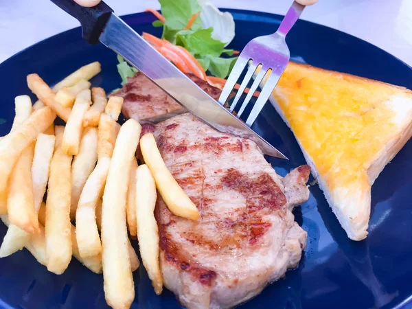 Ręce kobiety trzyma widelec i nóż do cięcia stek. To jest grillowany wieprzowina Chop Steak z sosu sos, są francuskie frytki i sałatka na bocznych potraw — Zdjęcie stockowe