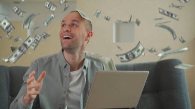 Başarılı genç adam dizüstü bilgisayarını kullanarak online iş kuruyor ve para basıyor. Dolarlar düşüyor..