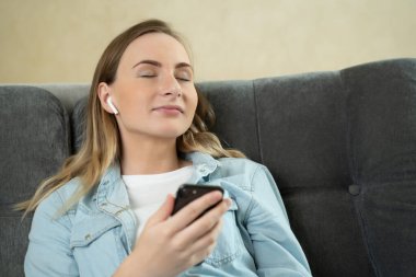 Genç bir kadın evdeki telefonunda kulaklıklarla müzik dinlerken kanepede gözleri kapalı rahatlıyor..