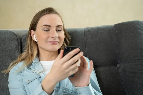 Mujer joven sentada escuchando música con auriculares en su teléfono móvil en casa mientras se relaja en un sofá . — Foto de Stock