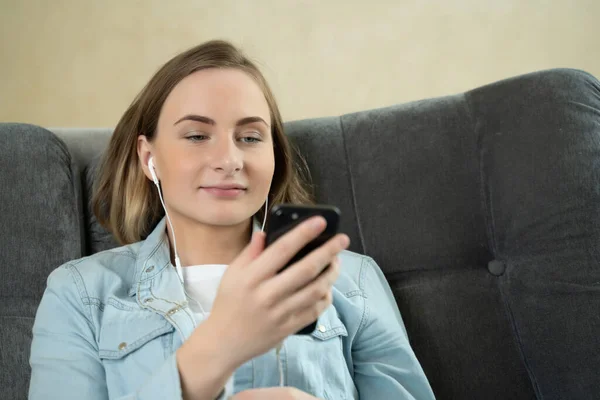Mujer joven sentada escuchando música con auriculares en su teléfono móvil en casa mientras se relaja en un sofá . — Foto de Stock
