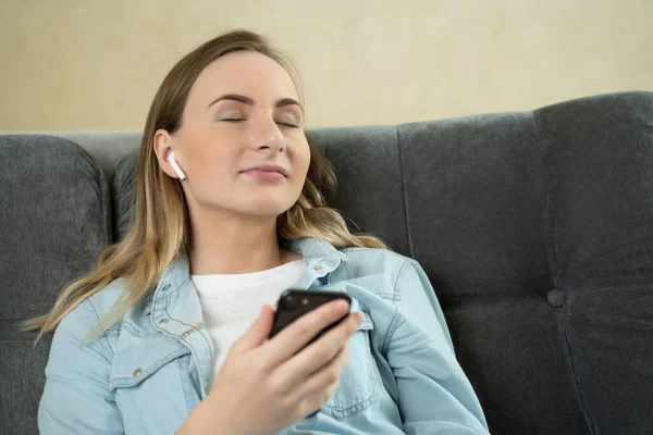 Mujer joven sentada escuchando música con auriculares en su teléfono móvil en casa mientras se relaja con los ojos cerrados en un sofá . — Foto de Stock