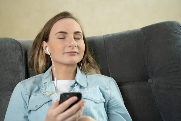 Jovem sentada ouvindo música com fones de ouvido em seu celular em casa enquanto relaxa com os olhos fechados em um sofá . — Fotografia de Stock