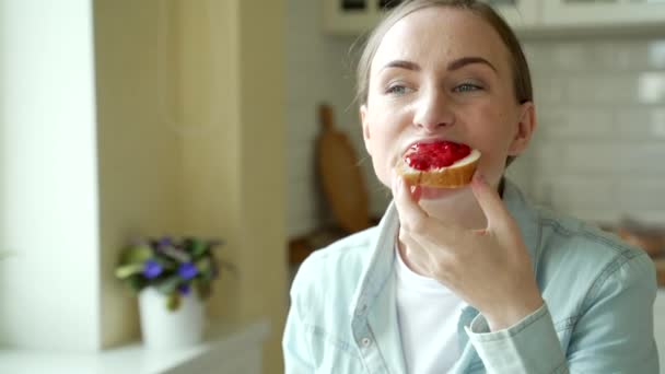 Mulher bonita comendo um delicioso sanduíche de geléia em casa na cozinha — Vídeo de Stock